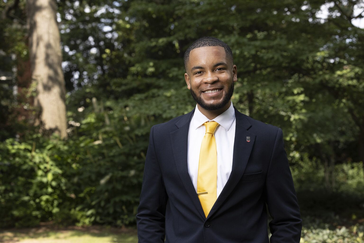 Eric Okanume, a Black student, smiles on campus in a dark suit with yellow tie and UGA pin.