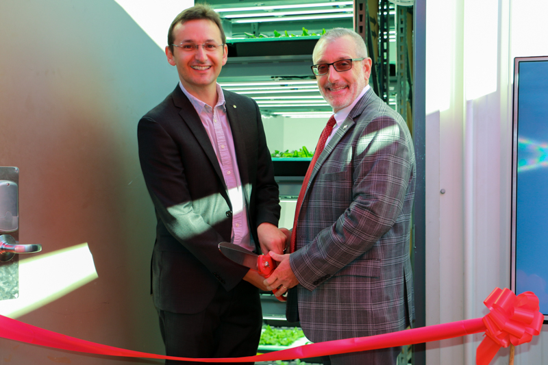 Two men in suits stand facing the camera, jointly holding a large pair of scissors in front of a red ribbon