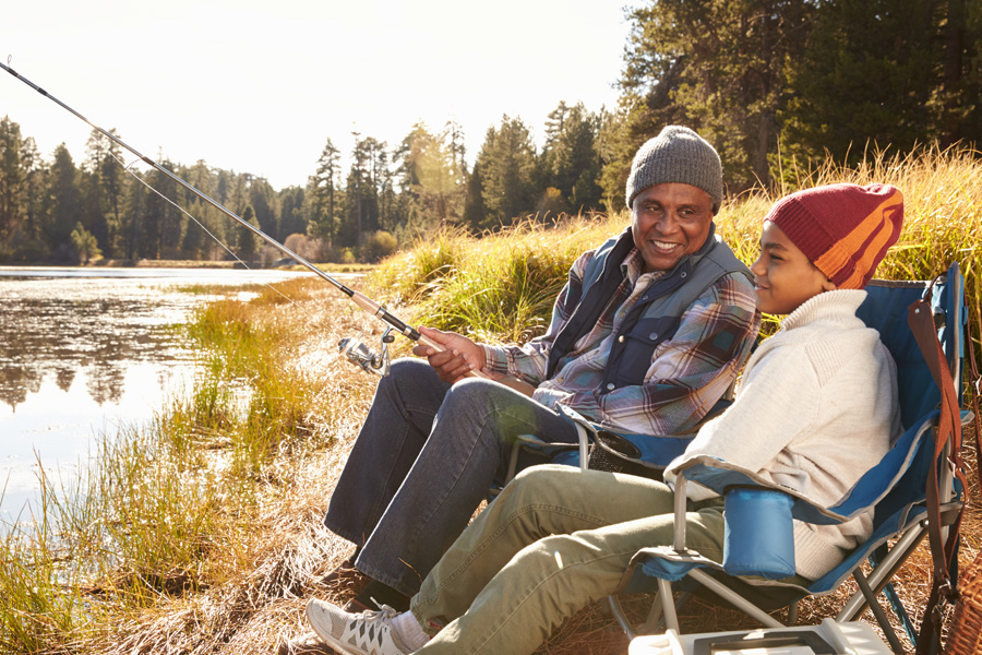 An older man and a young boy are sitting on the edge of a body of water and fishing while wearing clothing that suggests it is cold outside.