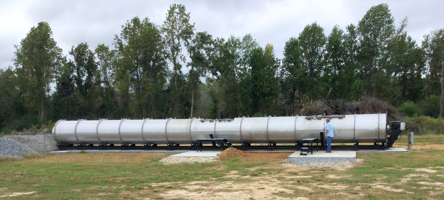 A drum composter, which looks like the tank on a tanker truck securely mounted onto a concrete slab, sits at the edge of a clearing on a farm.