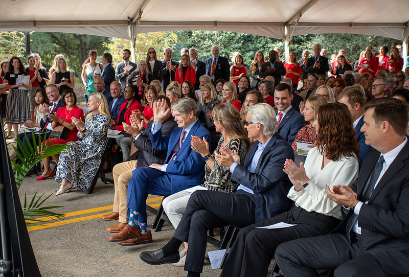 A man wearing a blue suit raises his hand in a crowd during a ribbon cutting ceremony.
