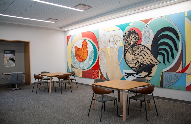 A student lounge with tables and chairs features brightly colored, wood-cut artwork of a hen and rooster