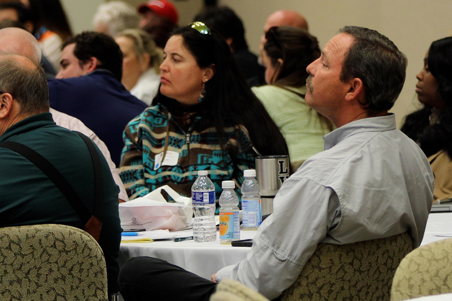 A group of conference attendees sit around a table, listening to a speaker.
