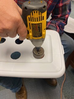 A hand-held drill bores holes into a plastic lid.