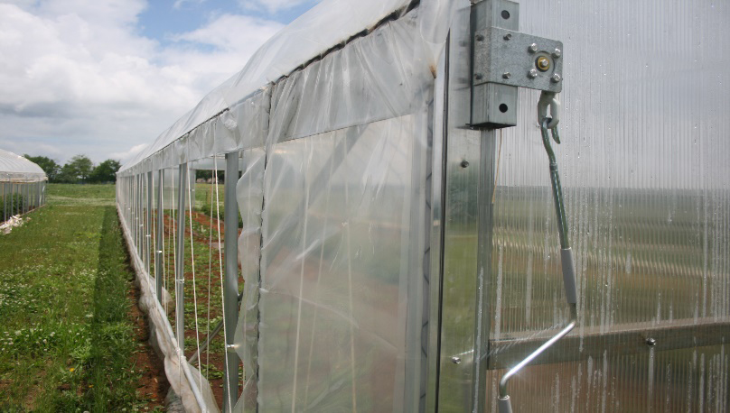 A view of a high-tunnel sidewall from the corner of the structure showing a crank used to raise or lower the sidewall plastic and the framing of the high tunnel.