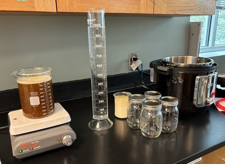 A nutrient broth sits on a counter in a flask next to a large graduated cylinder. There are also several jars and some YPD sitting next to a presure cooker.