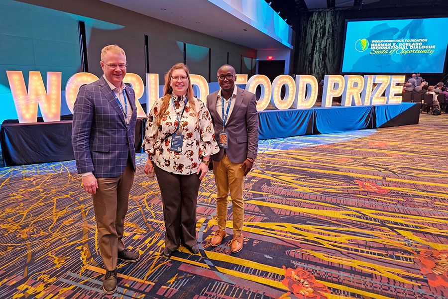 Three people stand together at an event for the World Food Prize, with large, illuminated letters spelling out "WORLD FOOD PRIZE" prominently displayed in the background. They are indoors in a spacious, well-lit venue with a colorful carpet pattern. Each person wears event lanyards and badges around their necks. The man on the left is dressed in a checkered blazer and beige pants, the woman in the middle wears a white blouse with a floral pattern and dark pants, and the man on the right sports a gray blazer, blue shirt, and tan pants. A large screen in the background shows the World Food Prize Foundation logo along with text that reads "Norman E. Borlaug // Seeds of Opportunity."