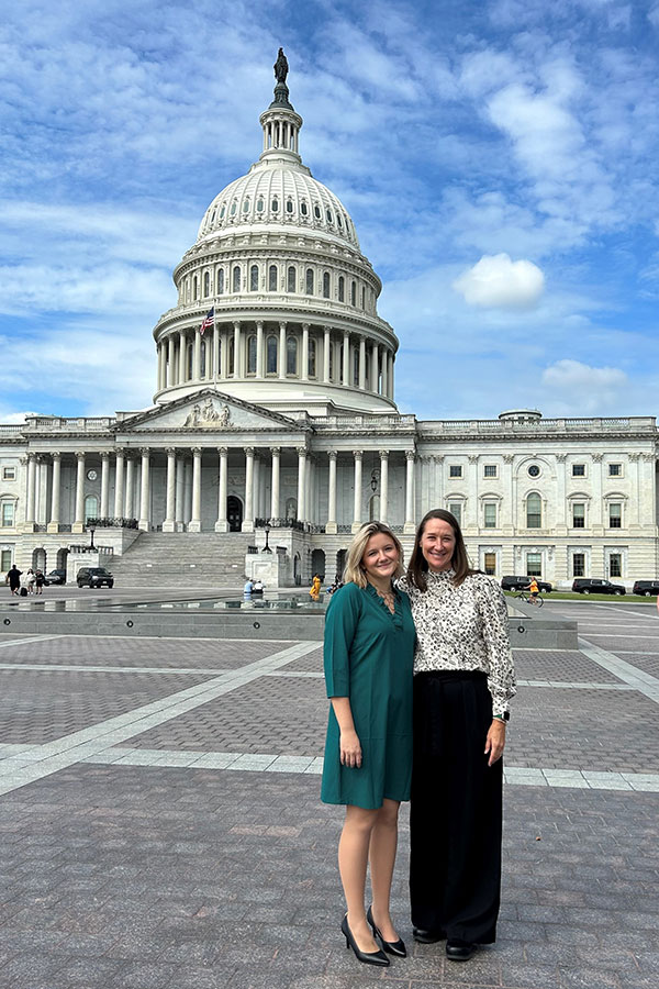 Two women, one with blonde hair wearing a green dress and the other a brunette wearing a black and white patterned shirt and black pants, stand with the U.S. Capitol in the background in Washington, D.C.