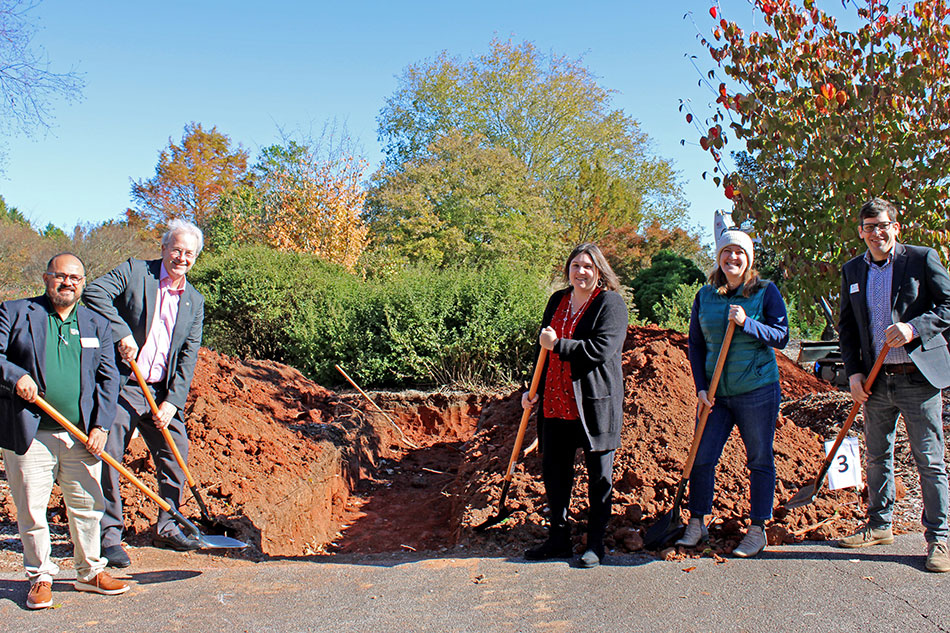 A group of five people holding shovels poses in front of a dug-out site, surrounded by piles of red dirt. The scene features vibrant autumn foliage, with colorful trees in the background. The individuals are participating in a groundbreaking ceremony, smiling and standing on paved ground.