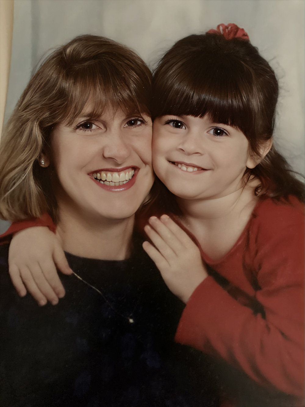 A mother and young daughter share a warm embrace, both smiling brightly, in a portrait with a soft, light background.