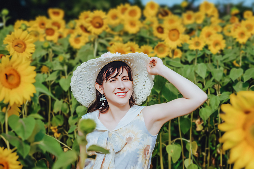 A smiling woman in a white sun hat and floral dress stands in a vibrant sunflower field.