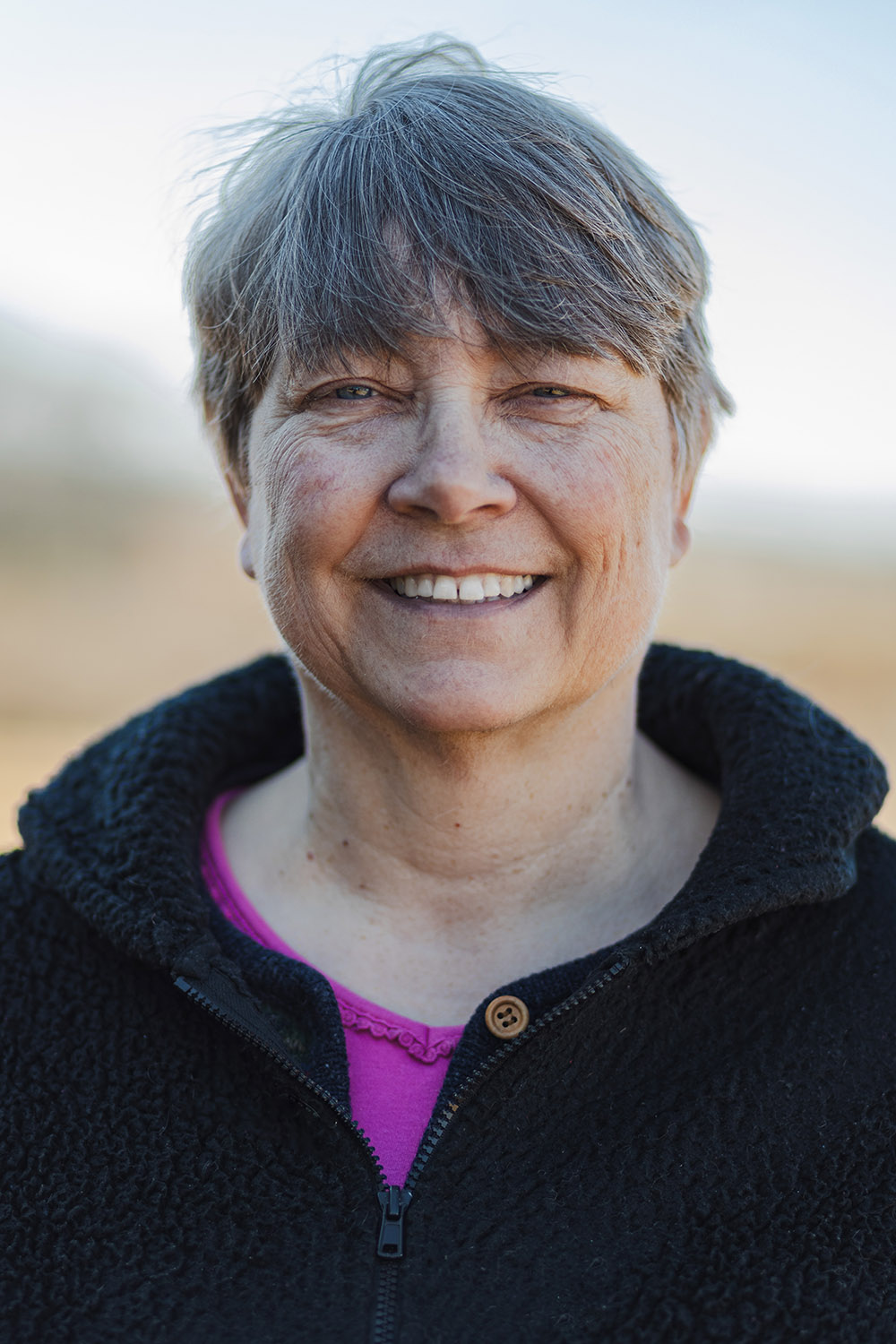 Smiling woman with short gray hair wearing a black jacket and pink top, standing outdoors with a blurred background.