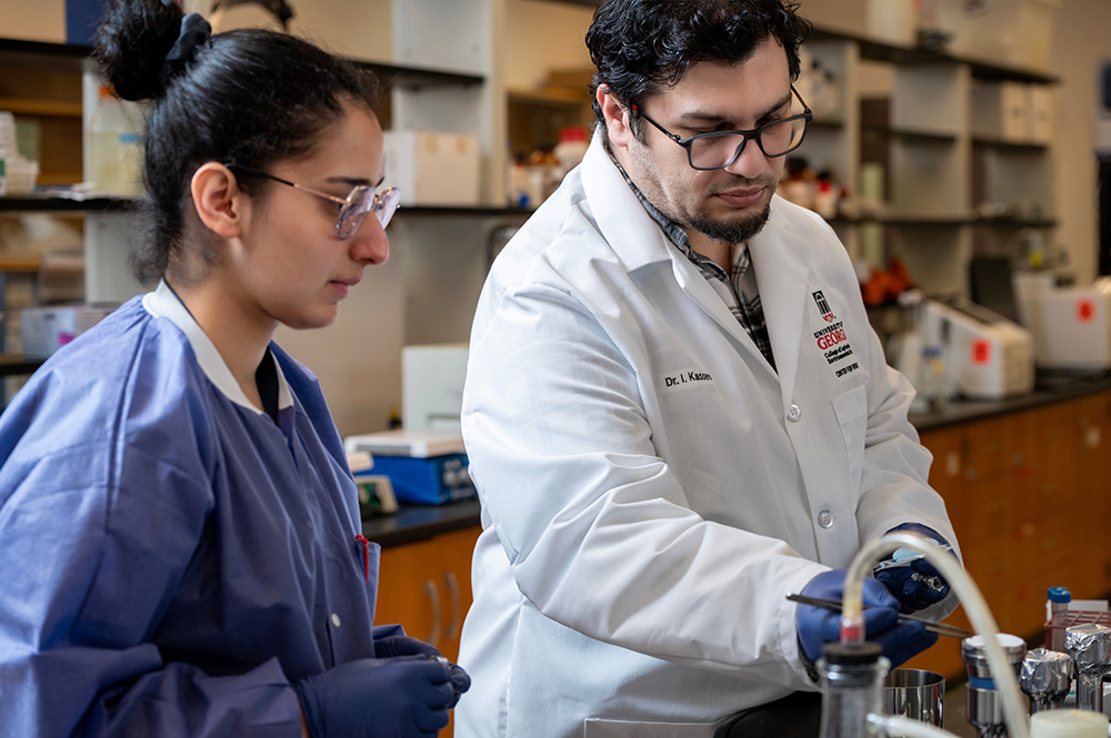 Two scientists in a lab, one in a white coat labeled 'Dr. I. Kassem,' working with equipment while the other observes.