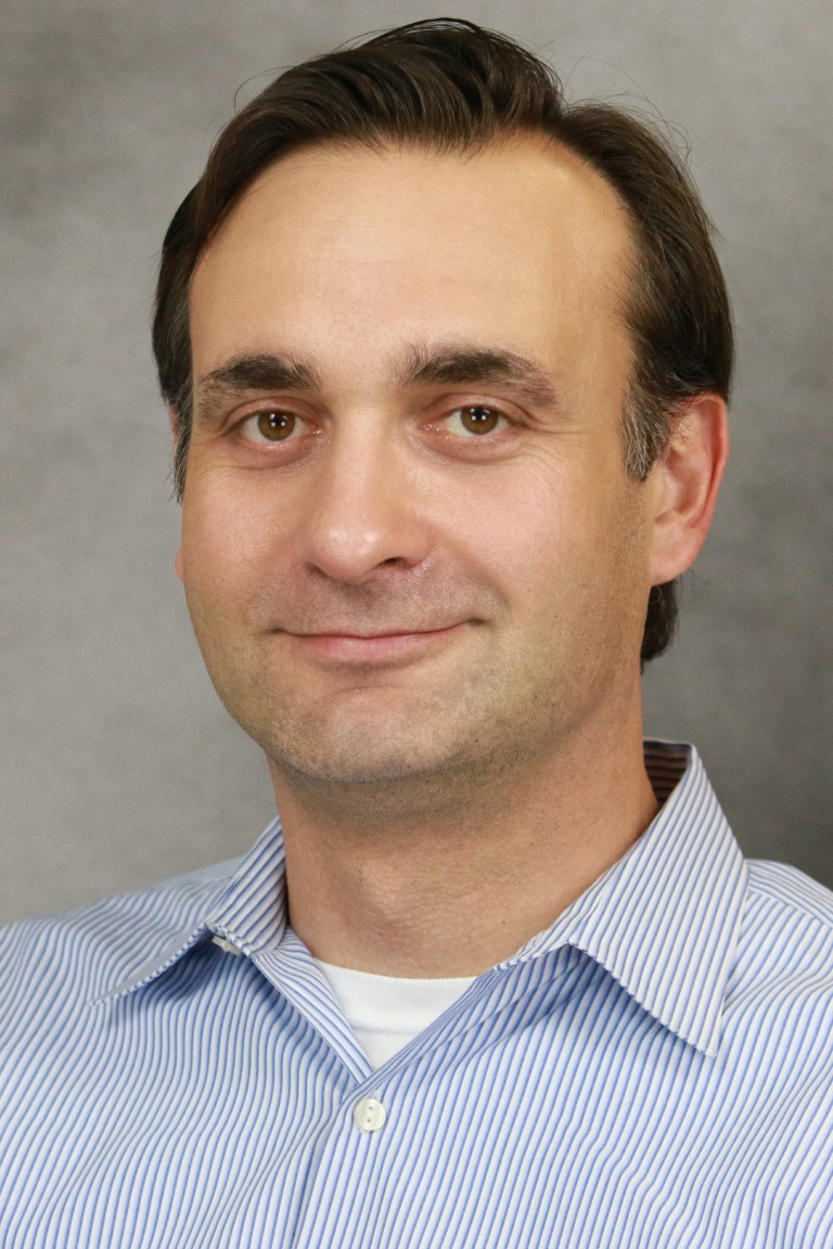 A photo of Michael Adjemian, a man with brown hair, brown eyes and a blue striped shirt.